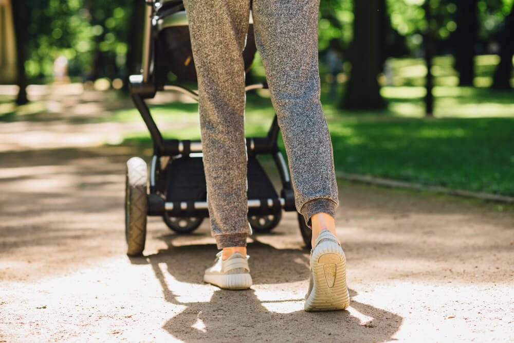 Young mother exercises in a park. Wears gray sport shoes, pushes a stroller.