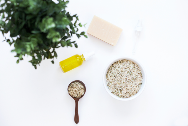 Person holding spoon full of hemp seeds with bottle of yellow oil and pink bar of soap in the background.