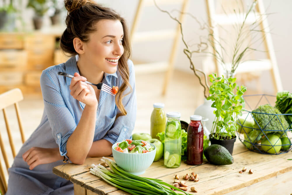 Woman in blue shirt eats salad surrounded by green vegetables and plants