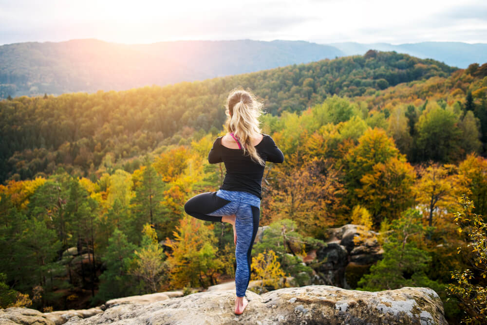Fall fitness woman in tree pose on a cliff with fall trees int he distance