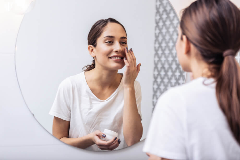 woman with dark hair putting on face cream in the mirror